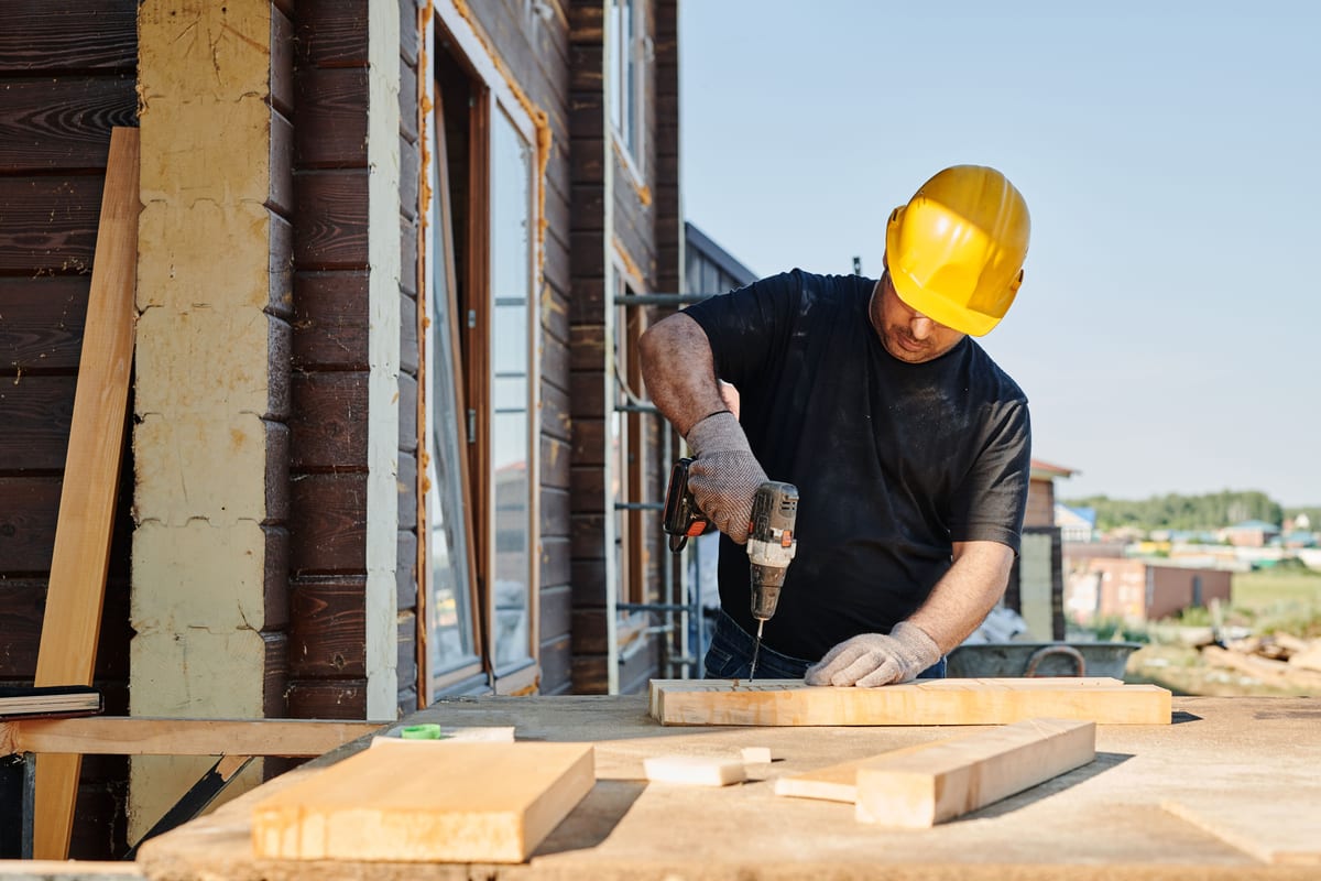 Contractor in a yellow hard hat and work gloves drilling into a board on a job site outside a wood-sided house