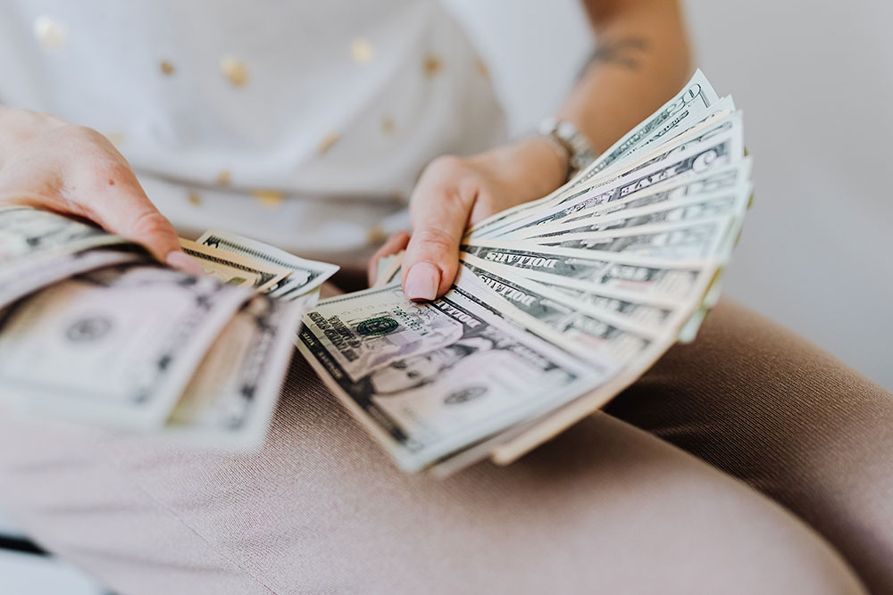 Close-up of a seated person fanning out a thick stack of US hundred-dollar bills across both hands, wearing a white top with small gold dots, a wristwatch, and dusty pink trousers