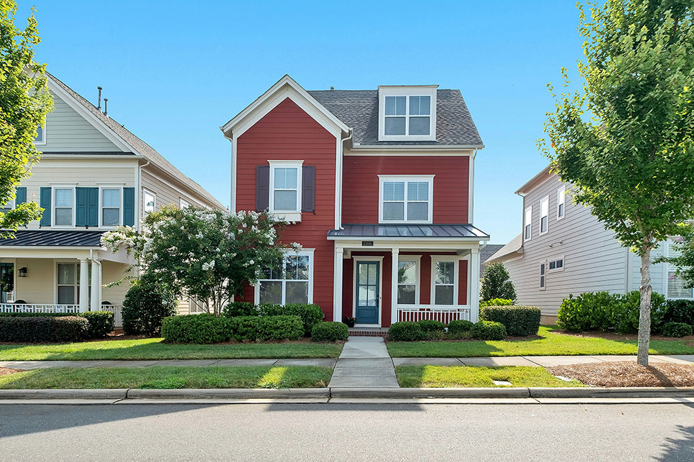 A red two-story house with white trim, dark shutters, a covered front porch, and a dormer window, set on a well-maintained lawn in a suburban neighborhood on a clear sunny day