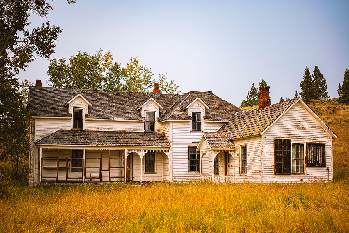 Abandoned white clapboard farmhouse with peeling paint, a mossy wood shingle roof, two brick chimneys, dormer windows, and a covered porch with decorative trim, standing in tall golden grass