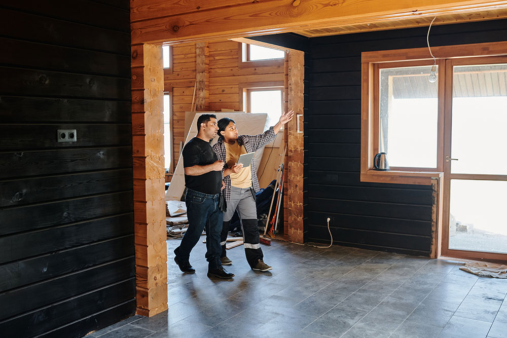 Two people in a room under renovation with dark timber walls, one in work trousers holding a tablet and pointing up