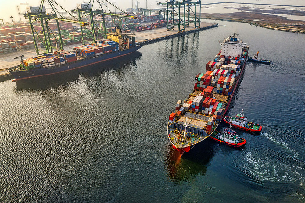 Aerial view of a container ship being guided by tugboats into a busy cargo port at dawn