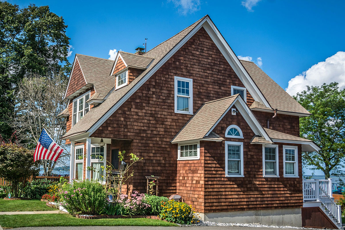Brown cedar-shingle home with white-trimmed double-hung windows and an arched gable window, an American flag flying by the front porch